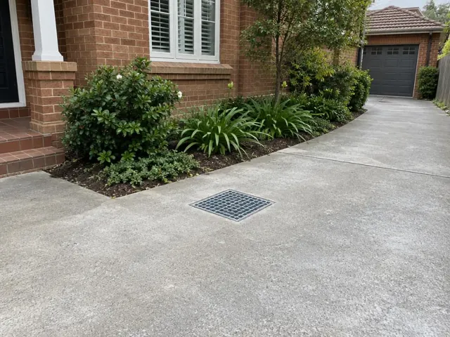 Concrete driveway with a square drain grate beside brick house and garden beds in Beverly Hills